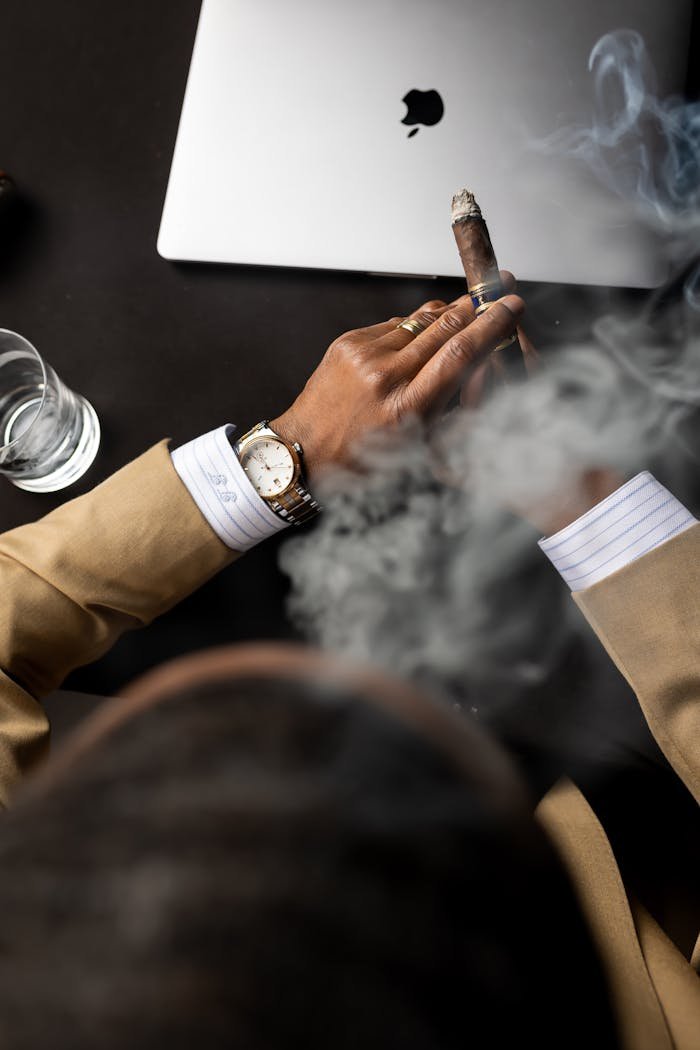 A businessman with a wristwatch holds a cigar beside a laptop, viewed from above in a smoky ambiance.